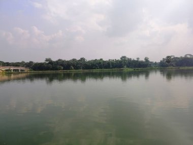 Yeşil ağaçlı sakin bir göl ve su yüzeyine yansıyan bulutlu bir gökyüzü. Net detaylar ve canlı renkler görsel cazibeyi artırır.