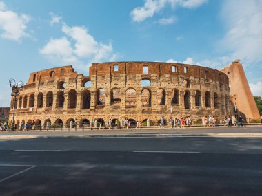 Roma 'daki tüm Kolezyum' un geniş panoramik sokak manzarası harap olmuş tuğla üst bölümü, çoklu kemer katları, güneşli mavi gökyüzünün altında yürüyen turistleri gösteriyor..