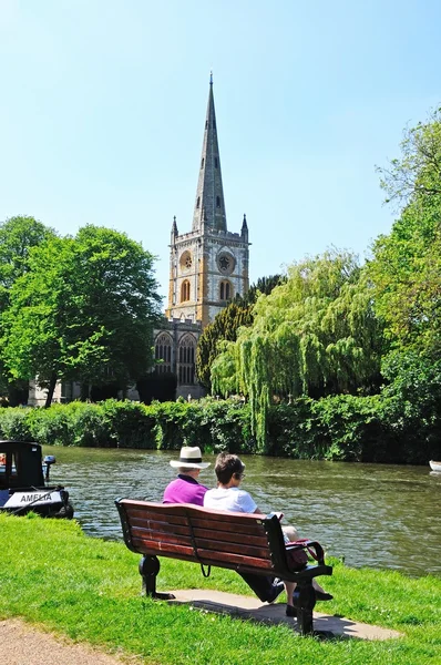 Holy Trinity Church seen across the River Avon with a couple sitting on a bench in the foreground, Stratford-Upon-Avon.