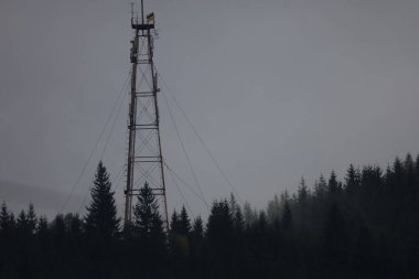 Ormandaki uzun metal telekom kulesi, kısmen sisle gizlenmiş. Ukrayna bayrağı tepede dalgalanıyor. Arka planda gri sisli gökyüzü.