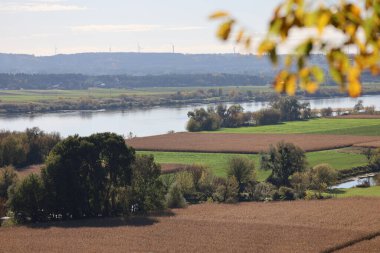 Geniş bir nehir, tarlalar ve tepeleri olan sonbahar kırsal arazisi. Orman ve birkaç rüzgar türbini parlak sisli bir gökyüzünün altında görülebilir..