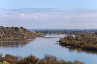 Gökyüzünü yansıtan sakin suyla Vistula Nehri 'nin geniş bir manzarası. Sarı ve yeşil sonbahar yaprakları her iki kıyıyı da kaplar, ortasında küçük bir kum çubuğu görünür.