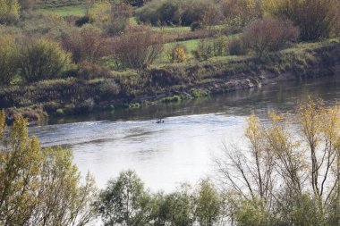 Sonbahar dalları tarafından çerçevelenmiş yumuşak bir akıntıya sahip geniş bir nehir. Uzak kıyılar, mevsimlik sarı ve kahverengi yapraklı dik kumlu katmanlar ve çalılar gösteriyor..