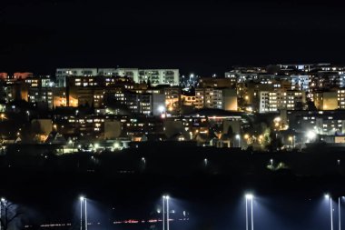 Geceleri aydınlık pencereler, ağaç siluetleri ve karanlık gökyüzünün altında aydınlık bir fabrika bacası olan Swiecie..