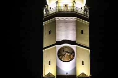 Sıcak ışıklarla aydınlatılan tarihi bir saat kulesinin detaylı bir görüntüsü, mimari özelliklerini gözler önüne seriyor ve en üstteki dekoratif korkulukları.