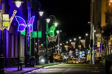 Gece bir şehir caddesi, büyük kelebek şekilleri ve ışık direkleri boyunca parıldayan yıldız motifleri de dahil olmak üzere canlı neon süslemelerle süslenir ve neşeli bir atmosfer yaratır.