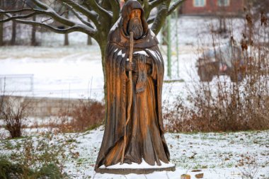 Yıpranmış ahşap bir heykel, elinde baston tutan cüppeli bir keşiş tasvir ediyor. Çıplak ağaçlar ve çalılarla karlı bir arazinin ortasında dikiliyor.