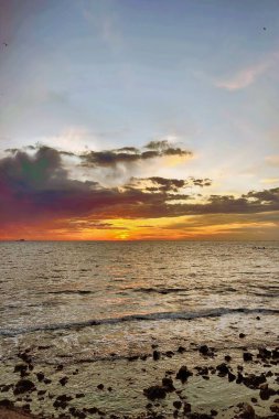 Ateşli Okyanus Günbatımı: Dramatik Gökyüzü, Rocky Shoreline.Yoğun, karamsar gün batımı canlı turuncu ve kırmızı bulutlar ve ön planda desenli, koyu kayalık bir plaj. Huzuru ve dramı yakalar.