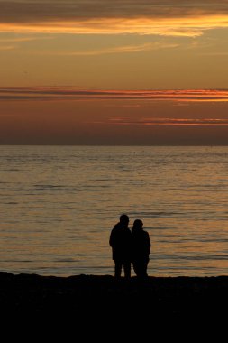 Sunset Beach 'teki Çiftin Silueti. Kumlu bir sahilde gölgede gölgeyle yakalanmış, altın bir denizin keyfini çıkaran bir çiftin huzurlu bir sahnesi. Güneş ufukta batarken iki insan arasında paylaşılan romantik ve sessiz bir an.