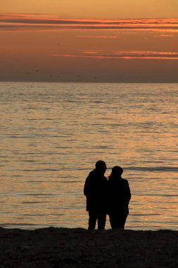 Sunset Beach 'teki Çiftin Silueti. Kumlu bir sahilde gölgede gölgeyle yakalanmış, altın bir denizin keyfini çıkaran bir çiftin huzurlu bir sahnesi. Güneş ufukta batarken iki insan arasında paylaşılan romantik ve sessiz bir an.