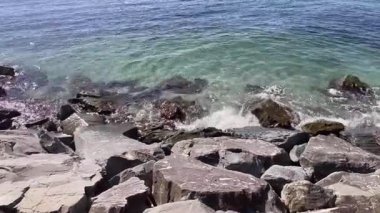 Rocky Shoreline 'a dalgalar çarpıyor. Açık turkuaz okyanus suyu güneşli bir günde koyu gri kıyı kayalarının üzerine hafifçe sıçrıyor. Doğal kayalık bir plajın yüksek açılı görüntüsü.