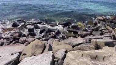 Rocky Shoreline 'a dalgalar çarpıyor. Açık turkuaz okyanus suyu güneşli bir günde koyu gri kıyı kayalarının üzerine hafifçe sıçrıyor. Doğal kayalık bir plajın yüksek açılı görüntüsü.
