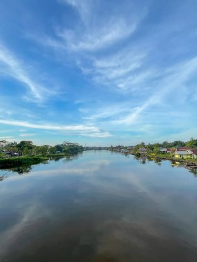Bereketli yeşil kıyılar arasında akan sakin bir nehir. Nehir kenarındaki evler. Geniş mavi gökyüzü altında yumuşak bulutlar..