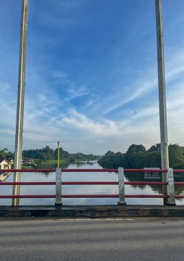 Yeşil kıyıları olan sakin bir nehre bakan bir köprüden ve yumuşak bulutlu geniş mavi gökyüzünün altında evler.