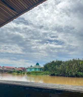 Çamurlu nehrin yanında yeşil kubbeli bir cami ve dramatik bulutlu bir gökyüzünün altında palmiye ağaçları..
