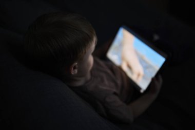 Üç yaşında bir çocuk, evinde bir kanepede oturup tablet ve akıllı telefondan çizgi film izliyor. Çocukluk kavramı, ekran zamanı, modern teknoloji ve boş zaman..