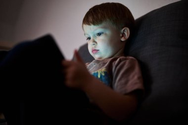 Üç yaşında bir çocuk, evinde bir kanepede oturup tablet ve akıllı telefondan çizgi film izliyor. Çocukluk kavramı, ekran zamanı, modern teknoloji ve boş zaman..