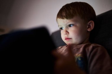 Üç yaşında bir çocuk, evinde bir kanepede oturup tablet ve akıllı telefondan çizgi film izliyor. Çocukluk kavramı, ekran zamanı, modern teknoloji ve boş zaman..