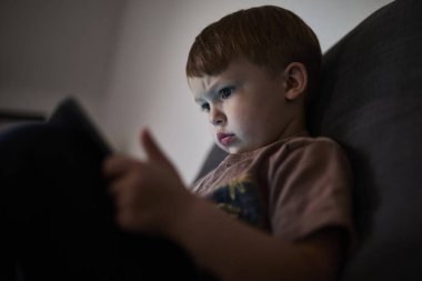 Üç yaşında bir çocuk, evinde bir kanepede oturup tablet ve akıllı telefondan çizgi film izliyor. Çocukluk kavramı, ekran zamanı, modern teknoloji ve boş zaman..