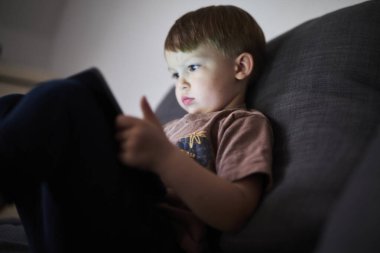 Üç yaşında bir çocuk, evinde bir kanepede oturup tablet ve akıllı telefondan çizgi film izliyor. Çocukluk kavramı, ekran zamanı, modern teknoloji ve boş zaman..