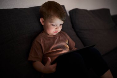 Üç yaşında bir çocuk, evinde bir kanepede oturup tablet ve akıllı telefondan çizgi film izliyor. Çocukluk kavramı, ekran zamanı, modern teknoloji ve boş zaman..