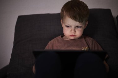 Üç yaşında bir çocuk, evinde bir kanepede oturup tablet ve akıllı telefondan çizgi film izliyor. Çocukluk kavramı, ekran zamanı, modern teknoloji ve boş zaman..