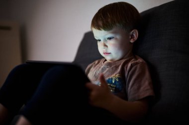 Üç yaşında bir çocuk, evinde bir kanepede oturup tablet ve akıllı telefondan çizgi film izliyor. Çocukluk kavramı, ekran zamanı, modern teknoloji ve boş zaman..
