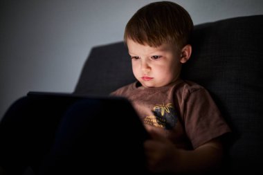 Üç yaşında bir çocuk, evinde bir kanepede oturup tablet ve akıllı telefondan çizgi film izliyor. Çocukluk kavramı, ekran zamanı, modern teknoloji ve boş zaman..