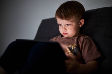 Üç yaşında bir çocuk, evinde bir kanepede oturup tablet ve akıllı telefondan çizgi film izliyor. Çocukluk kavramı, ekran zamanı, modern teknoloji ve boş zaman..
