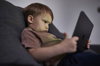 Üç yaşında bir çocuk, evinde bir kanepede oturup tablet ve akıllı telefondan çizgi film izliyor. Çocukluk kavramı, ekran zamanı, modern teknoloji ve boş zaman..