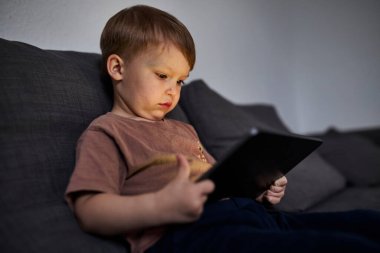 Üç yaşında bir çocuk, evinde bir kanepede oturup tablet ve akıllı telefondan çizgi film izliyor. Çocukluk kavramı, ekran zamanı, modern teknoloji ve boş zaman..