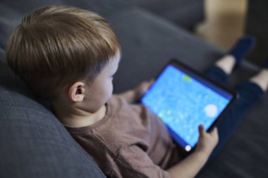 Üç yaşında bir çocuk, evinde bir kanepede oturup tablet ve akıllı telefondan çizgi film izliyor. Çocukluk kavramı, ekran zamanı, modern teknoloji ve boş zaman..