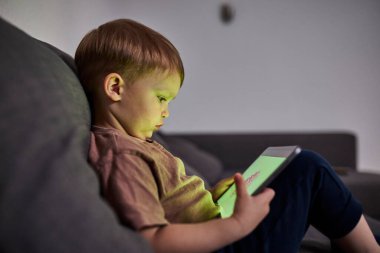 Üç yaşında bir çocuk, evinde bir kanepede oturup tablet ve akıllı telefondan çizgi film izliyor. Çocukluk kavramı, ekran zamanı, modern teknoloji ve boş zaman..