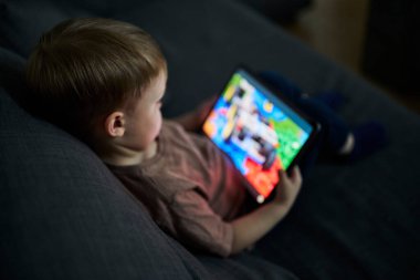 Üç yaşında bir çocuk, evinde bir kanepede oturup tablet ve akıllı telefondan çizgi film izliyor. Çocukluk kavramı, ekran zamanı, modern teknoloji ve boş zaman..