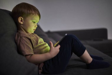 Üç yaşında bir çocuk, evinde bir kanepede oturup tablet ve akıllı telefondan çizgi film izliyor. Çocukluk kavramı, ekran zamanı, modern teknoloji ve boş zaman..