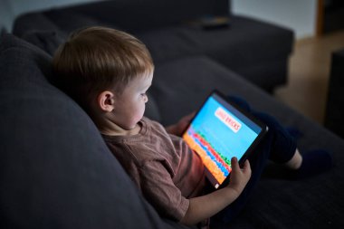 Üç yaşında bir çocuk, evinde bir kanepede oturup tablet ve akıllı telefondan çizgi film izliyor. Çocukluk kavramı, ekran zamanı, modern teknoloji ve boş zaman..