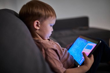 Üç yaşında bir çocuk, evinde bir kanepede oturup tablet ve akıllı telefondan çizgi film izliyor. Çocukluk kavramı, ekran zamanı, modern teknoloji ve boş zaman..