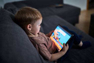 Üç yaşında bir çocuk, evinde bir kanepede oturup tablet ve akıllı telefondan çizgi film izliyor. Çocukluk kavramı, ekran zamanı, modern teknoloji ve boş zaman..