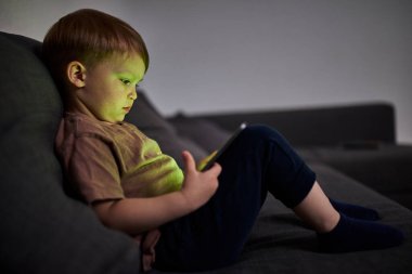 Üç yaşında bir çocuk, evinde bir kanepede oturup tablet ve akıllı telefondan çizgi film izliyor. Çocukluk kavramı, ekran zamanı, modern teknoloji ve boş zaman..