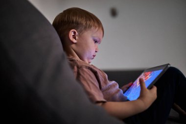Üç yaşında bir çocuk, evinde bir kanepede oturup tablet ve akıllı telefondan çizgi film izliyor. Çocukluk kavramı, ekran zamanı, modern teknoloji ve boş zaman..