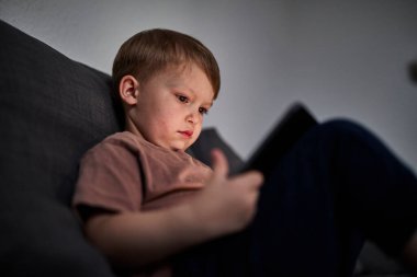Üç yaşında bir çocuk, evinde bir kanepede oturup tablet ve akıllı telefondan çizgi film izliyor. Çocukluk kavramı, ekran zamanı, modern teknoloji ve boş zaman..