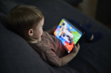 Üç yaşında bir çocuk, evinde bir kanepede oturup tablet ve akıllı telefondan çizgi film izliyor. Çocukluk kavramı, ekran zamanı, modern teknoloji ve boş zaman..