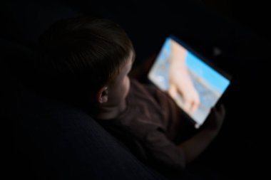 Üç yaşında bir çocuk, evinde bir kanepede oturup tablet ve akıllı telefondan çizgi film izliyor. Çocukluk kavramı, ekran zamanı, modern teknoloji ve boş zaman..