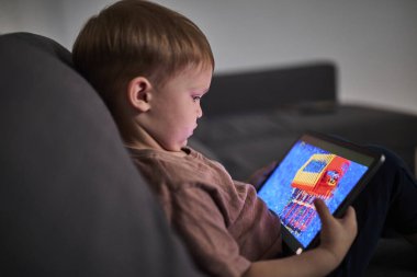 Üç yaşında bir çocuk, evinde bir kanepede oturup tablet ve akıllı telefondan çizgi film izliyor. Çocukluk kavramı, ekran zamanı, modern teknoloji ve boş zaman..
