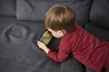 Üç yaşında bir çocuk, evinde bir kanepede oturup tablet ve akıllı telefondan çizgi film izliyor. Çocukluk kavramı, ekran zamanı, modern teknoloji ve boş zaman..