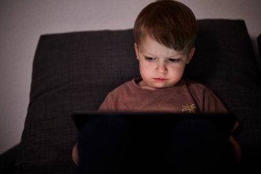 Üç yaşında bir çocuk, evinde bir kanepede oturup tablet ve akıllı telefondan çizgi film izliyor. Çocukluk kavramı, ekran zamanı, modern teknoloji ve boş zaman..