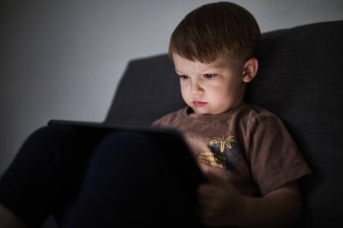 Üç yaşında bir çocuk, evinde bir kanepede oturup tablet ve akıllı telefondan çizgi film izliyor. Çocukluk kavramı, ekran zamanı, modern teknoloji ve boş zaman..