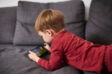 Üç yaşında bir çocuk, evinde bir kanepede oturup tablet ve akıllı telefondan çizgi film izliyor. Çocukluk kavramı, ekran zamanı, modern teknoloji ve boş zaman..