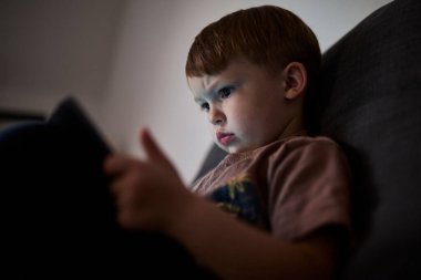 Üç yaşında bir çocuk, evinde bir kanepede oturup tablet ve akıllı telefondan çizgi film izliyor. Çocukluk kavramı, ekran zamanı, modern teknoloji ve boş zaman..