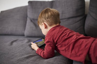Üç yaşında bir çocuk, evinde bir kanepede oturup tablet ve akıllı telefondan çizgi film izliyor. Çocukluk kavramı, ekran zamanı, modern teknoloji ve boş zaman..